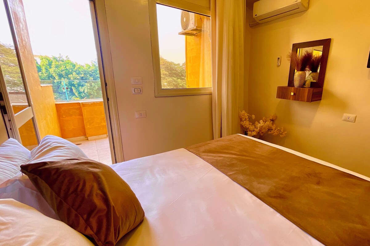 A hotel room with a bed, a TV, and a window with a view of greenery.