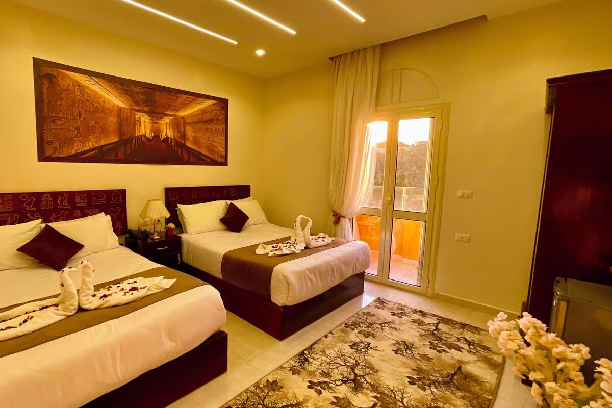 A hotel room with two beds, a large wall mural, a balcony with curtains, and a floral arrangement.