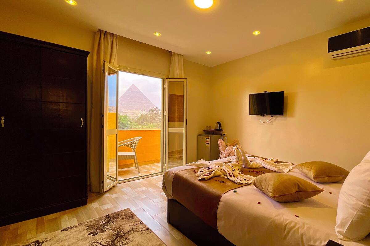 A bedroom with a bed, a TV, a minibar, and a view of a pyramid.