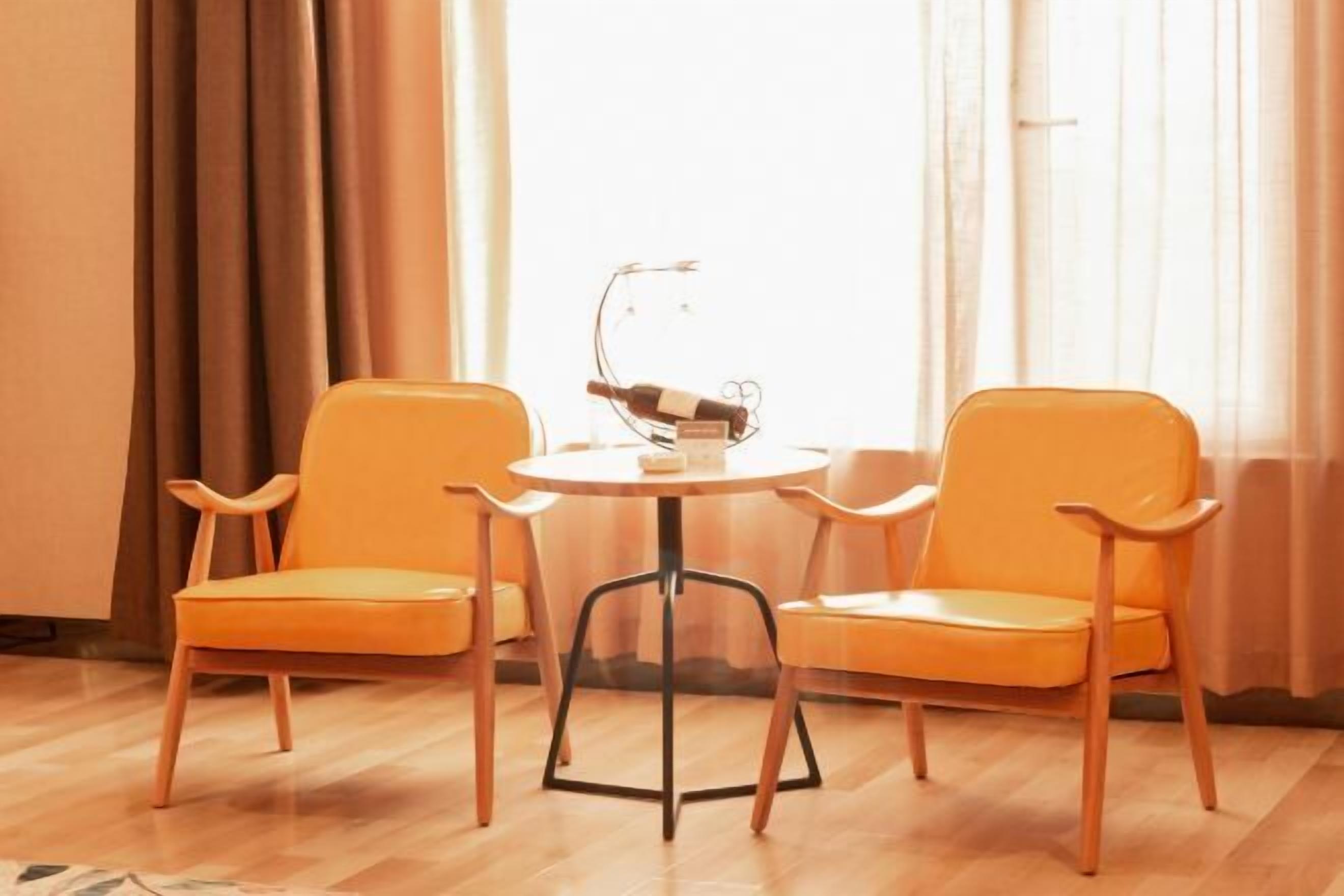 A room with two orange chairs and a small round table, a bottle of wine on a stand, and a window with curtains.