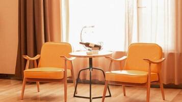 A room with two orange chairs and a small round table, a bottle of wine on a stand, and a window with curtains.