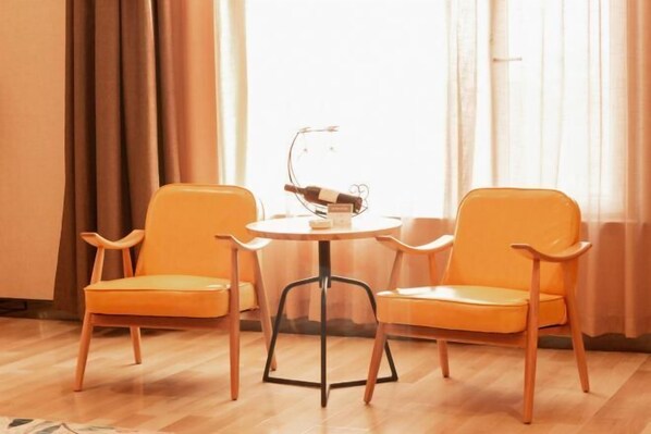 Une pièce avec deux fauteuils orange et une petite table ronde, une bouteille de vin posée sur un présentoir, et une fenêtre avec des rideaux.