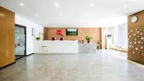 A hotel reception area with a reception desk, two receptionists, a television screen, and a wall with a clock and notes. - Shankee Tea Style Hotel (Qingdao Jiaodong International Airport Haier Avenue) (Jiaozhou)