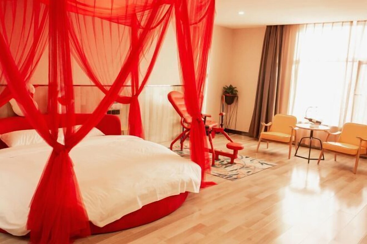 Une chambre d’hôtel moderne avec un lit à baldaquin rouge, un pouf rond rouge, une petite table avec des chaises et une vue sur la ville grâce à de grandes fenêtres.