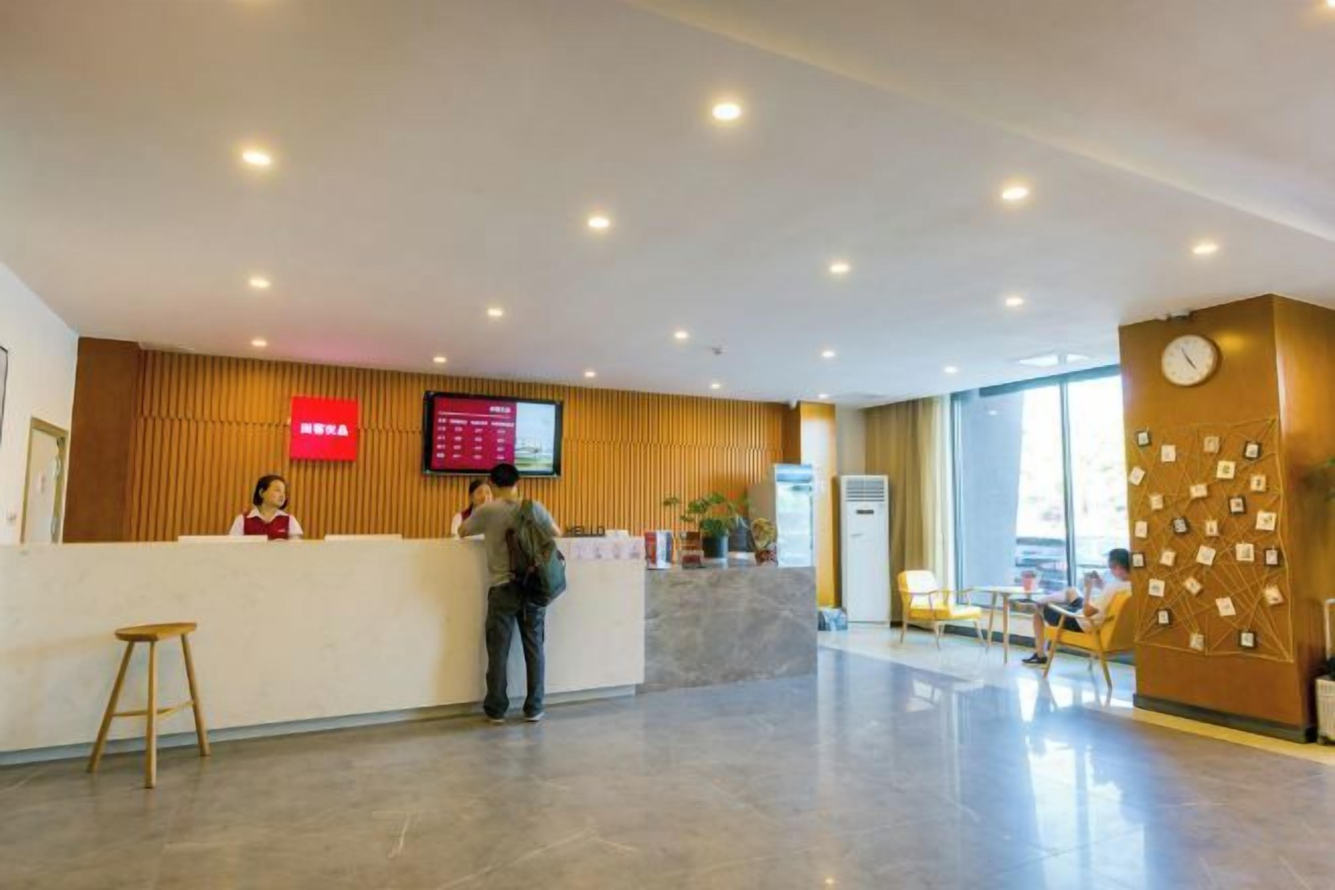 Área de recepção de hotel, com balcão, tela digital e espaço de estar com mesas e cadeiras.