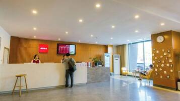 Área de recepção de hotel, com balcão, tela digital e espaço de estar com mesas e cadeiras.