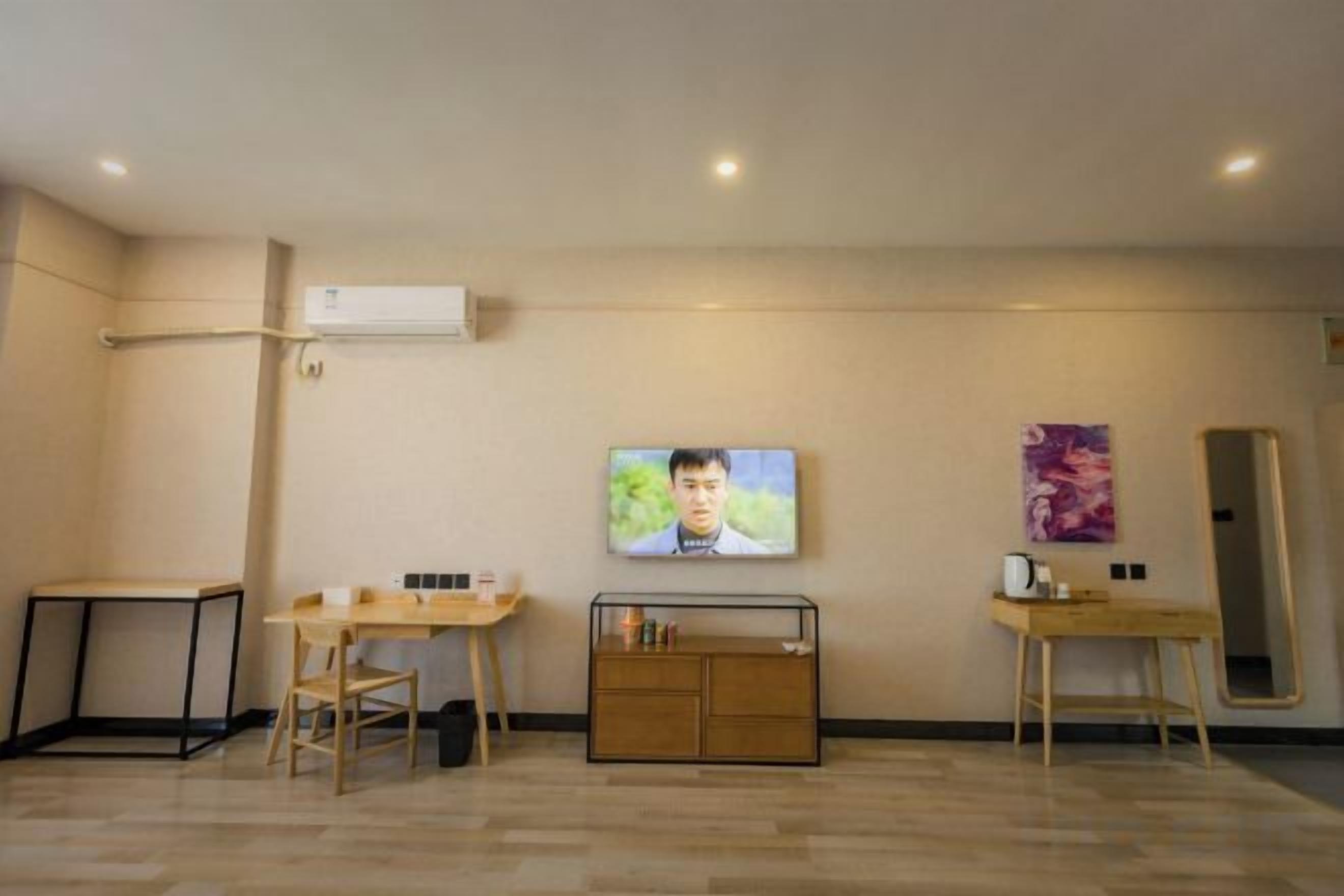 A hotel room with a flat-screen TV, a wooden desk, a chair, a small cabinet, and a mirror on the wall.
