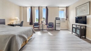 Ett hotellrum med en säng, ett skrivbord med en lampa, två stolar, en tv och ett kylskåp.