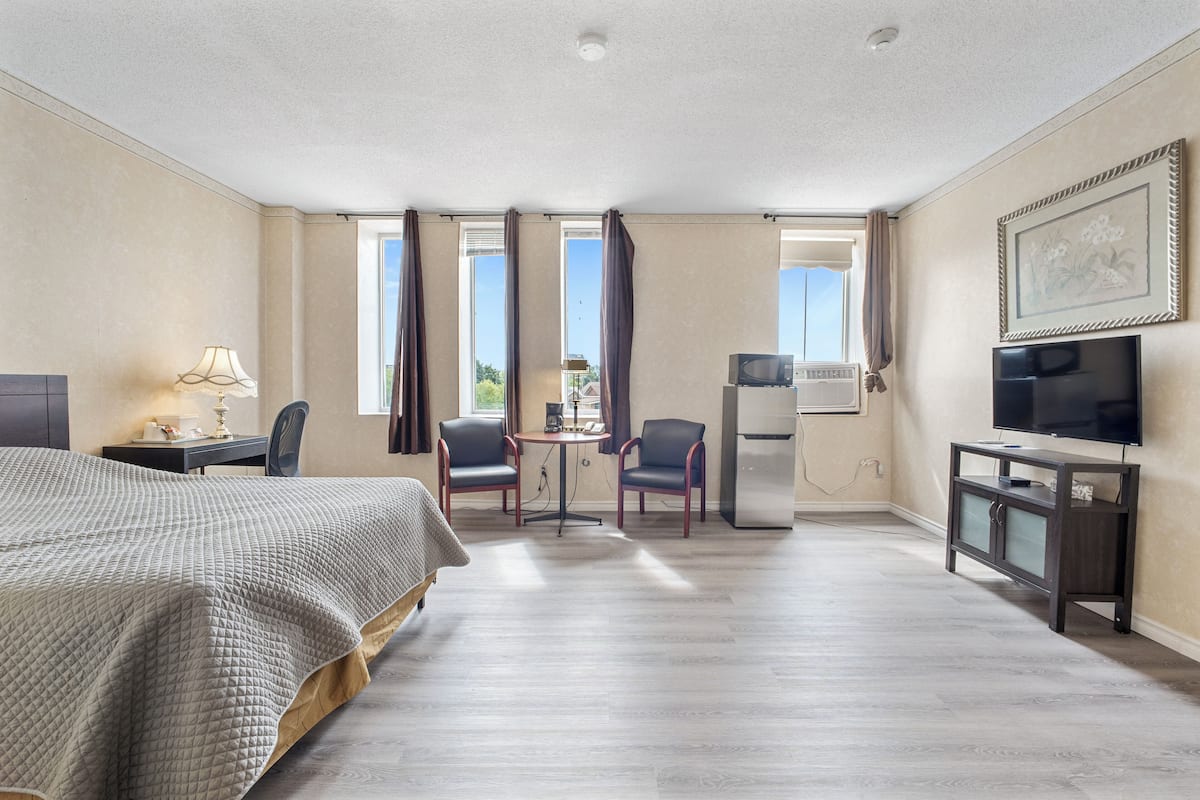 A hotel room with a bed, a desk with a lamp, two chairs, a television, and a refrigerator.