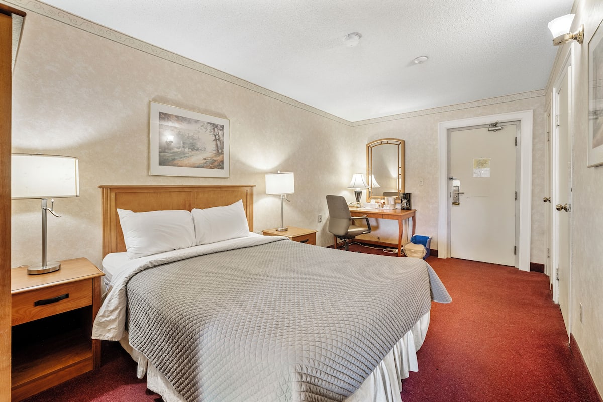 A hotel room with a large bed, bedside tables with lamps, a desk with a chair, a mirror, and a door.