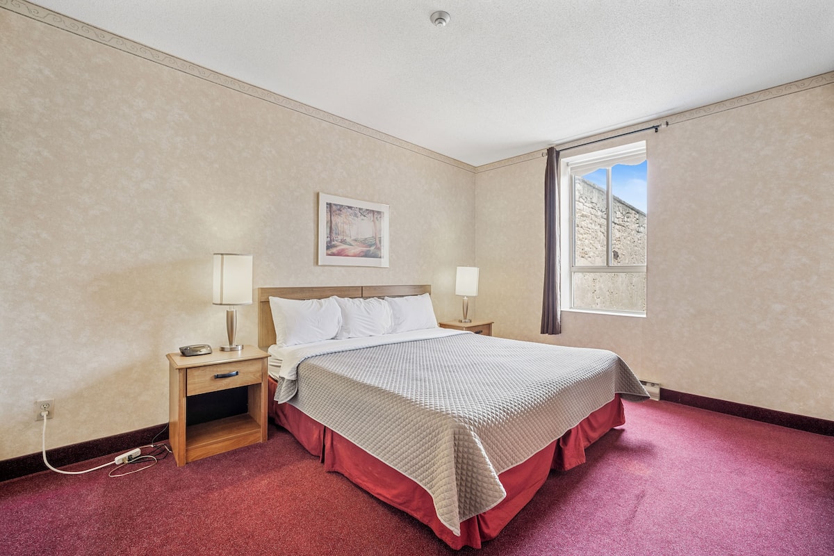 A hotel room with a bed, two bedside tables with lamps, a painting on the wall, and a window with curtains.
