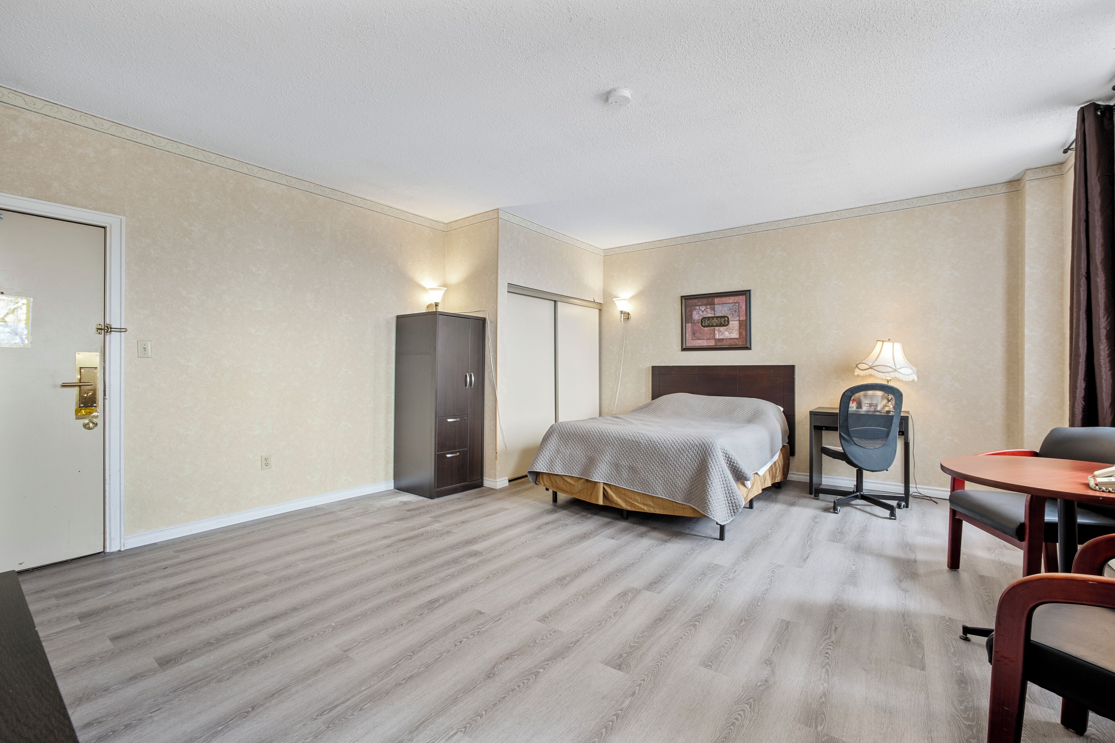 A hotel room with a bed, a desk with a chair, a wardrobe, and a lamp.