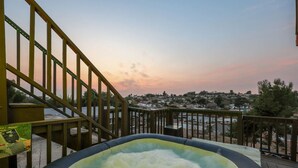 Banheira de hidromassagem em deck de madeira, com vista para uma área residencial e o pôr do sol.