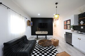 A modern living room with a black sofa, a wooden coffee table, a foosball table, and a kitchen area with white cabinets and a black backsplash.