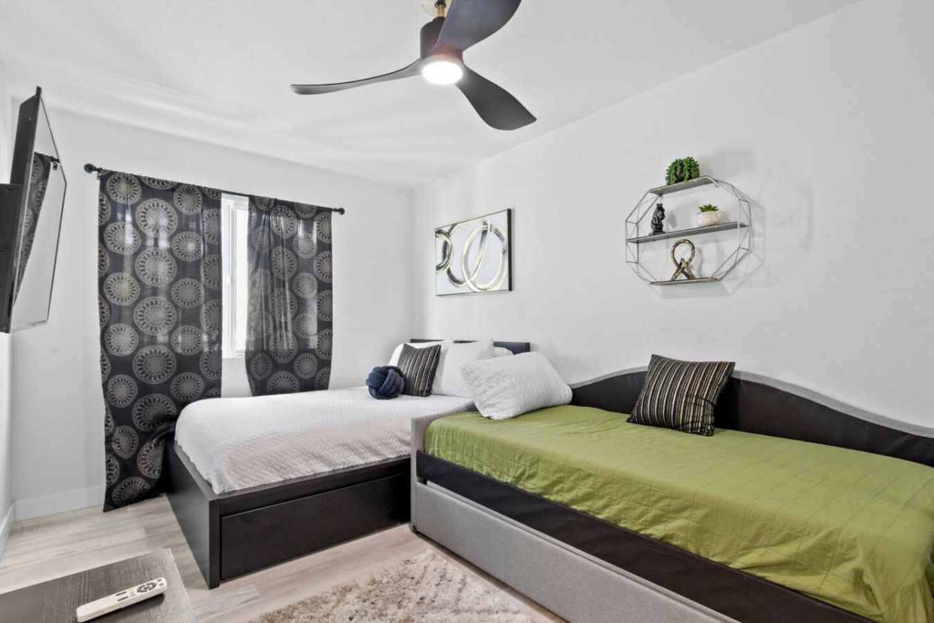 Quarto com duas camas, ventoinha no teto, prateleira na parede com objetos decorativos e janela com cortinas estampadas.