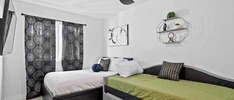 Ein Schlafzimmer mit zwei Betten, einem Deckenventilator, einem an der Wand befestigten Regal mit Dekoration und einem Fenster mit gemusterten Vorhängen.