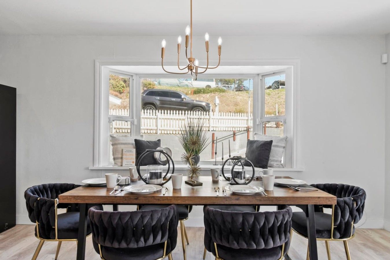 A dining area with a wooden table, black upholstered chairs, a chandelier, and a view of a car through a large window.