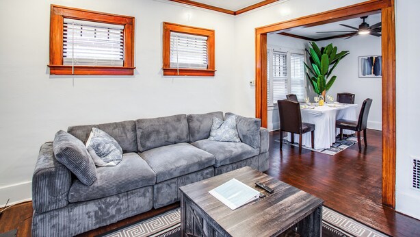 A living room with a grey sectional sofa, a wooden coffee table, and two windows with wooden frames.