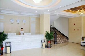 A hotel reception area with a receptionist, a reception desk, a staircase, and potted plants.
