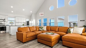 Une cuisine moderne avec des meubles blancs et des appareils en acier inoxydable, un grand canapé d’angle en forme de L avec une table d’appoint, et un coin repas avec une table et des chaises.