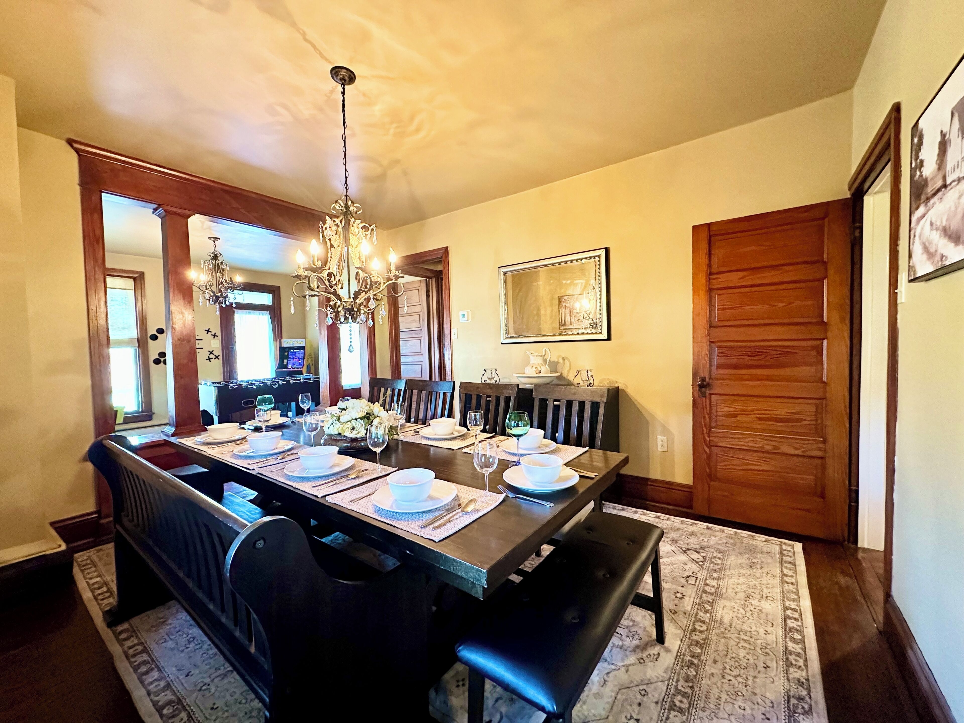 Une salle à manger avec une longue table dressée pour six personnes, un lustre, des chaises en bois et un banc.