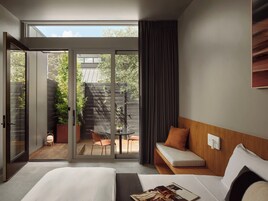 Une chambre d’hôtel moderne dotée d’un grand lit, d’un coin salon avec un banc rembourré, d’une petite table et d’une porte-fenêtre coulissante donnant sur un patio extérieur avec une terrasse en bois et des plantes en pot.