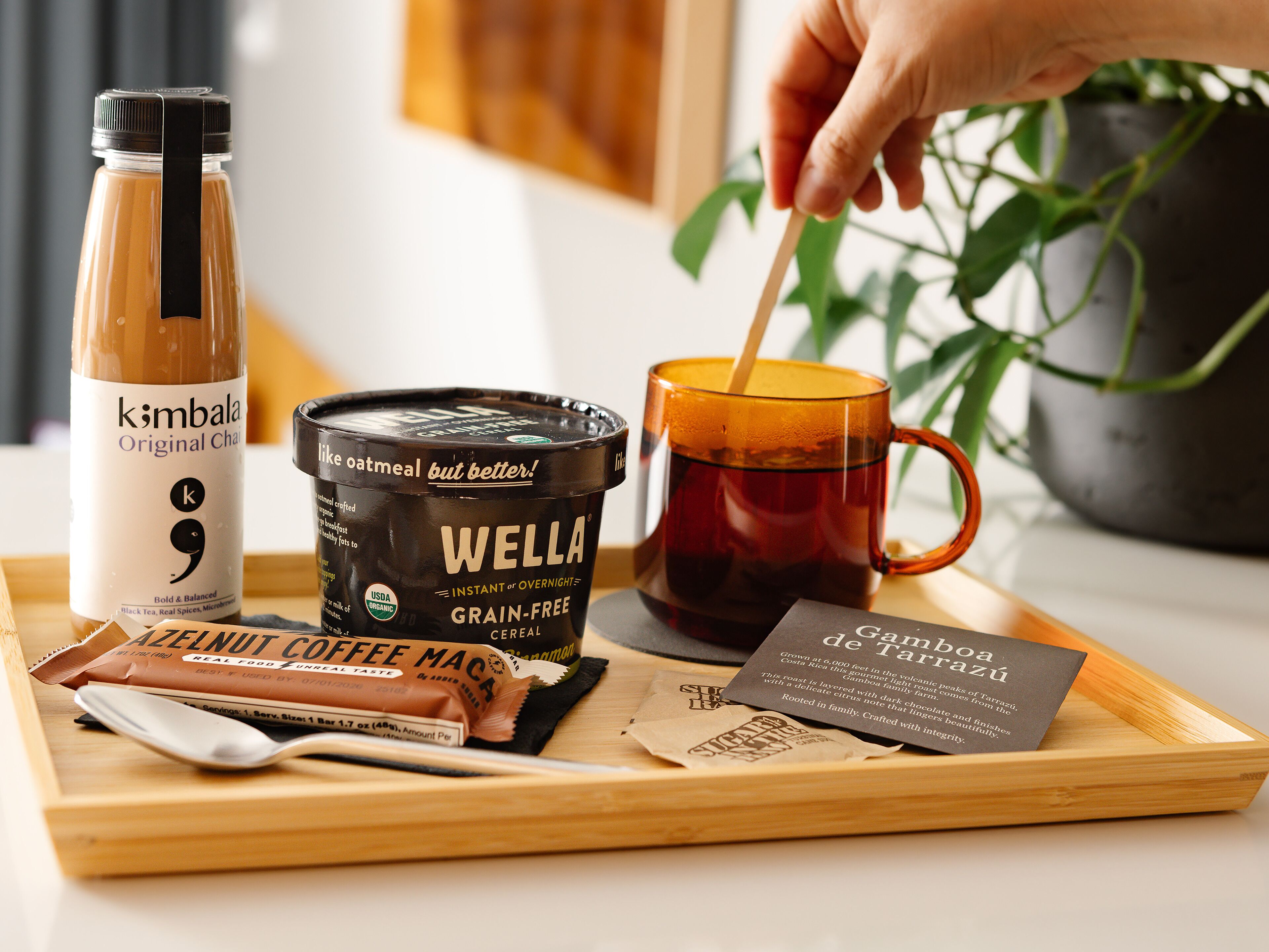 A tray with a bottle of kombucha, a container of Wella grain-free cereal, a mug of tea, and a packet of coffee macchiato.