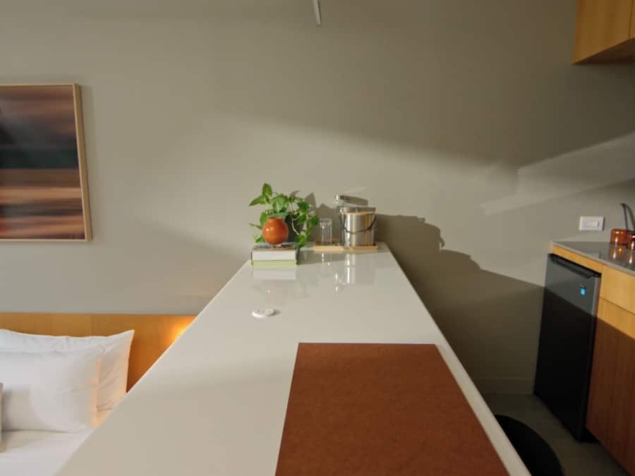 A modern hotel room with a large bed, a bedside table, a small kitchen area, and a large window.