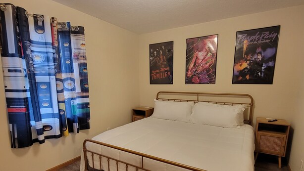 A bedroom with a metal-framed bed, white bedding, wooden flooring, and three posters on the wall. - RiverTown Retro | King Bed | Near Deaconess | Pet-Friendly (Newburgh)