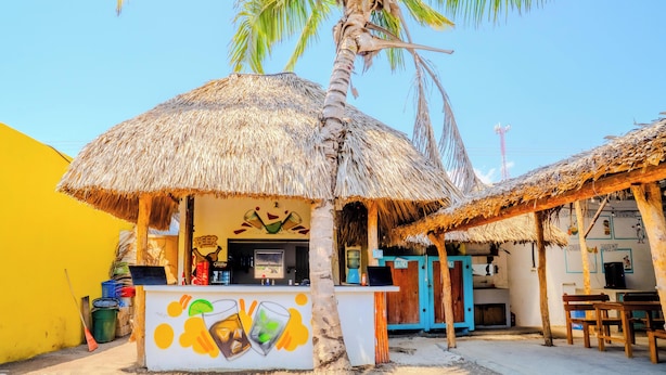 Un pequeño bar al aire libre con techo de paja, pared amarilla y mostrador con carta de bebidas.