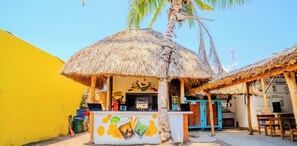 Un pequeño bar al aire libre con techo de paja, pared amarilla y mostrador con carta de bebidas.