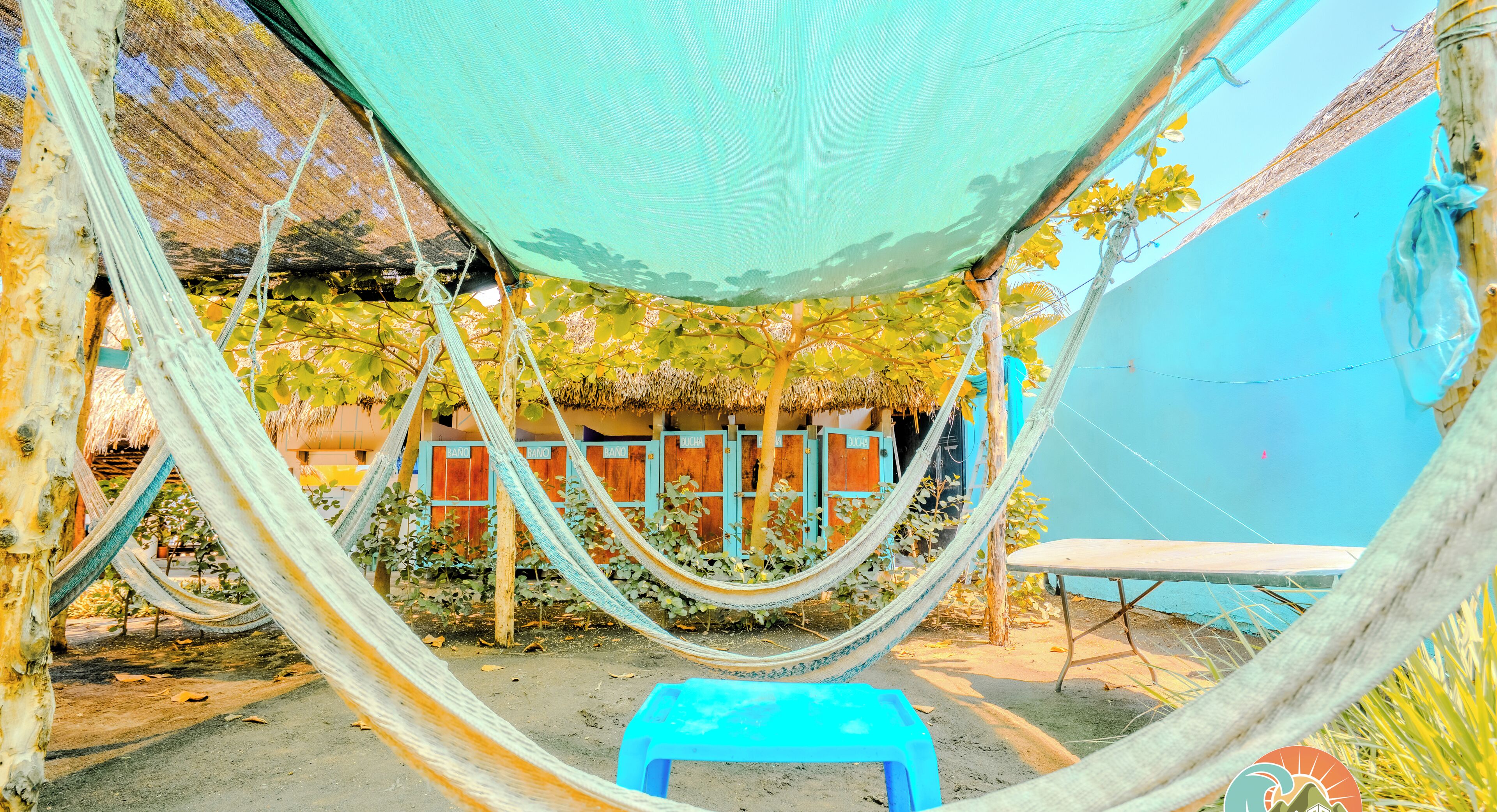 A hammock setup with a blue plastic stool, a thatched-roof structure, and a blue wall in the background.