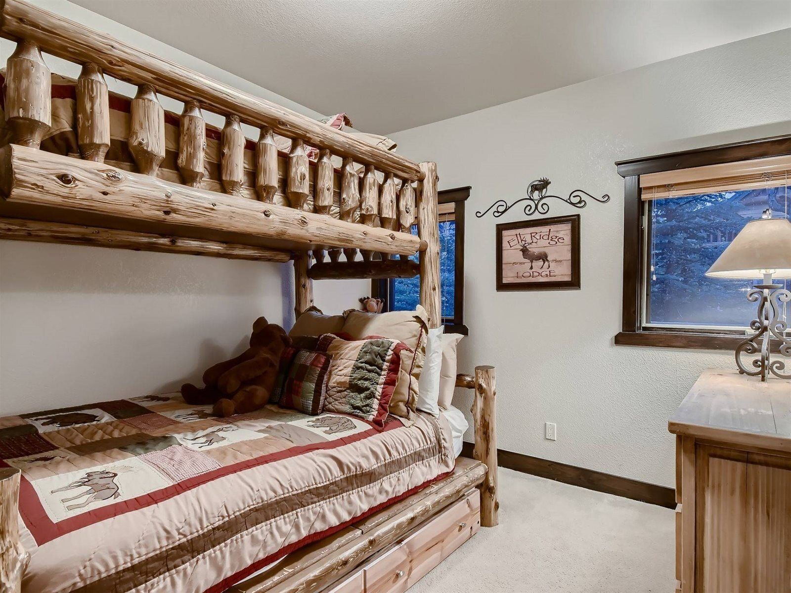 A rustic bedroom with a wooden bunk bed, a quilted bedspread, a framed picture on the wall, and a window with a view of trees.