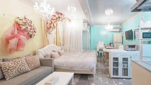 A bedroom with a bed, a sofa, a bedside table, a chandelier, and a kitchen area with cabinets and appliances. - Sakura (Nagoya)