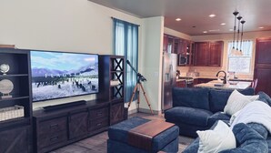 Een moderne woonkamer met een flatscreen-televisie, donker houten meubels, een blauwe bank en een keuken met houten kastjes en een roestvrijstalen koelkast.