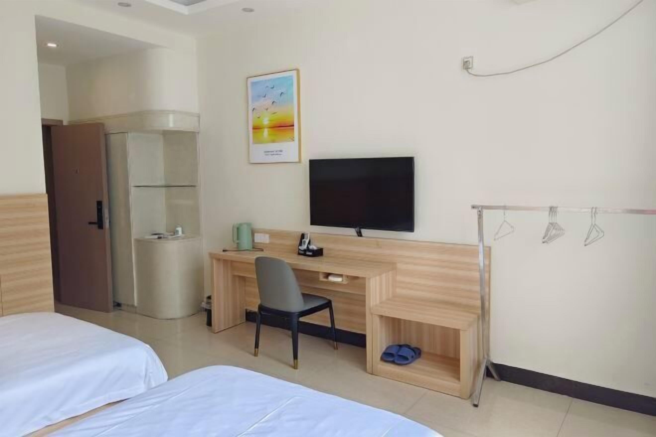 A hotel room with two beds, a desk with a chair, a television, a small shelf, and a shoe rack.