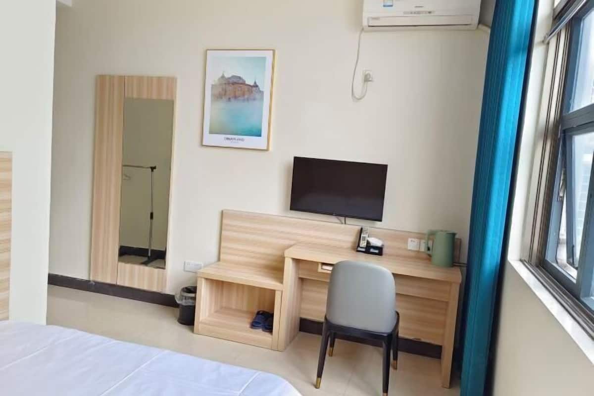 A hotel room with a bed, a desk with a chair, a television, a framed picture, and a window with curtains.