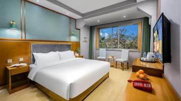 Habitación de hotel con una cama grande, una mesita de noche con teléfono, una pequeña mesa con un frutero, un televisor en la pared y una ventana con vista a árboles.