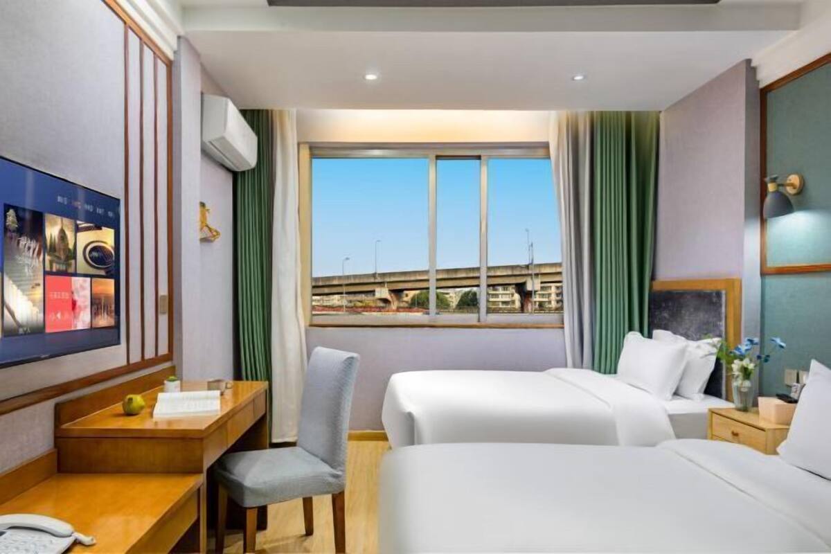 A hotel room with two beds, a desk, a chair, a TV, and a window with a view of buildings.