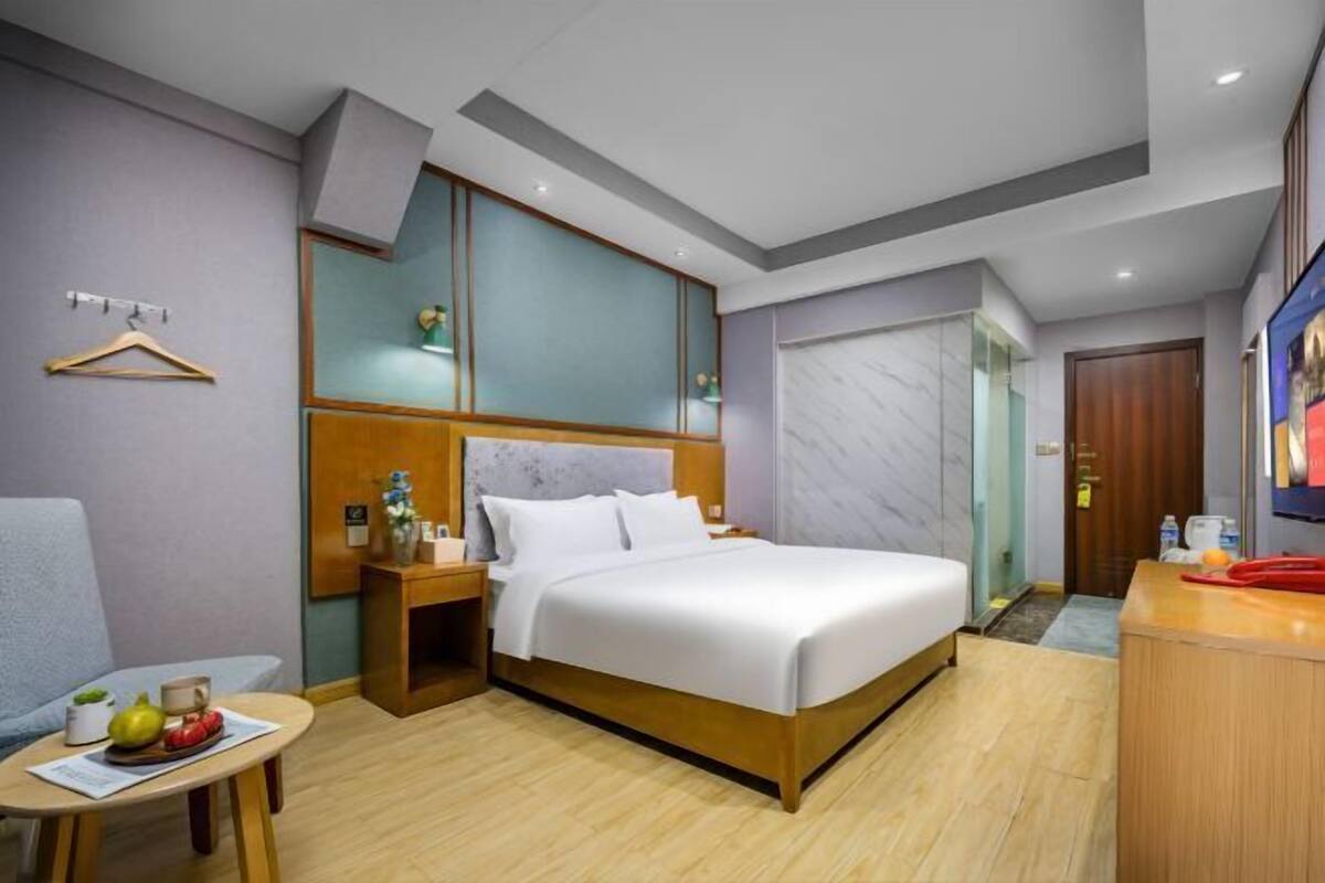 A hotel room with a bed, bedside table, chair, and a small table with fruits and a cup.