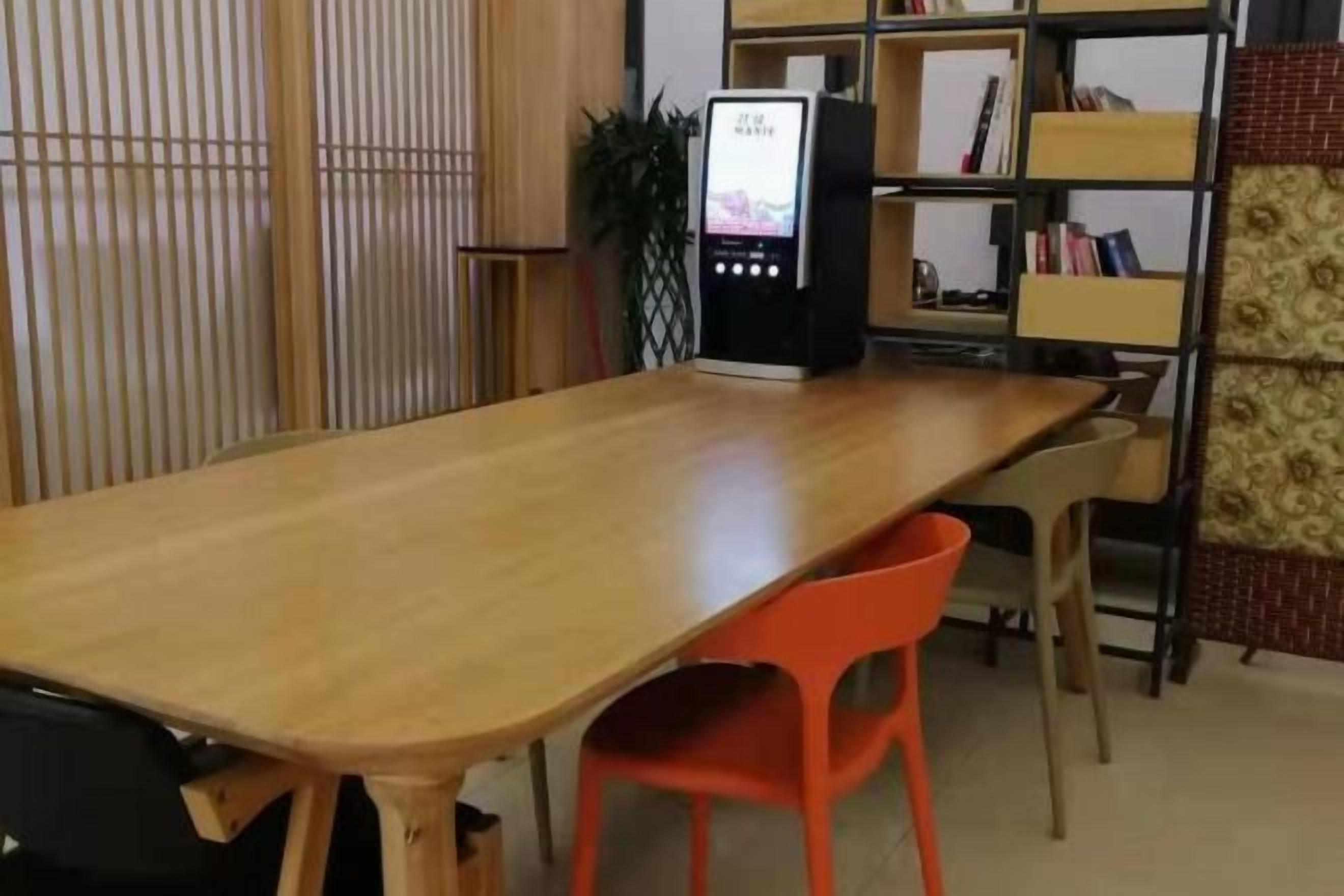 Una mesa de madera con dos sillas anaranjadas, una máquina expendedora y una estantería en una habitación con pared estampada.