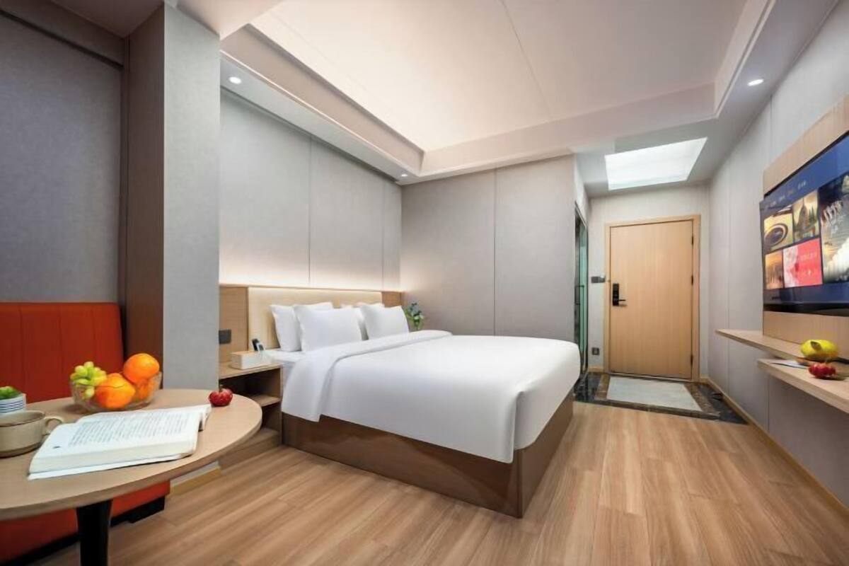 A modern hotel room with a bed, a desk with a laptop, a fruit bowl, and a door.