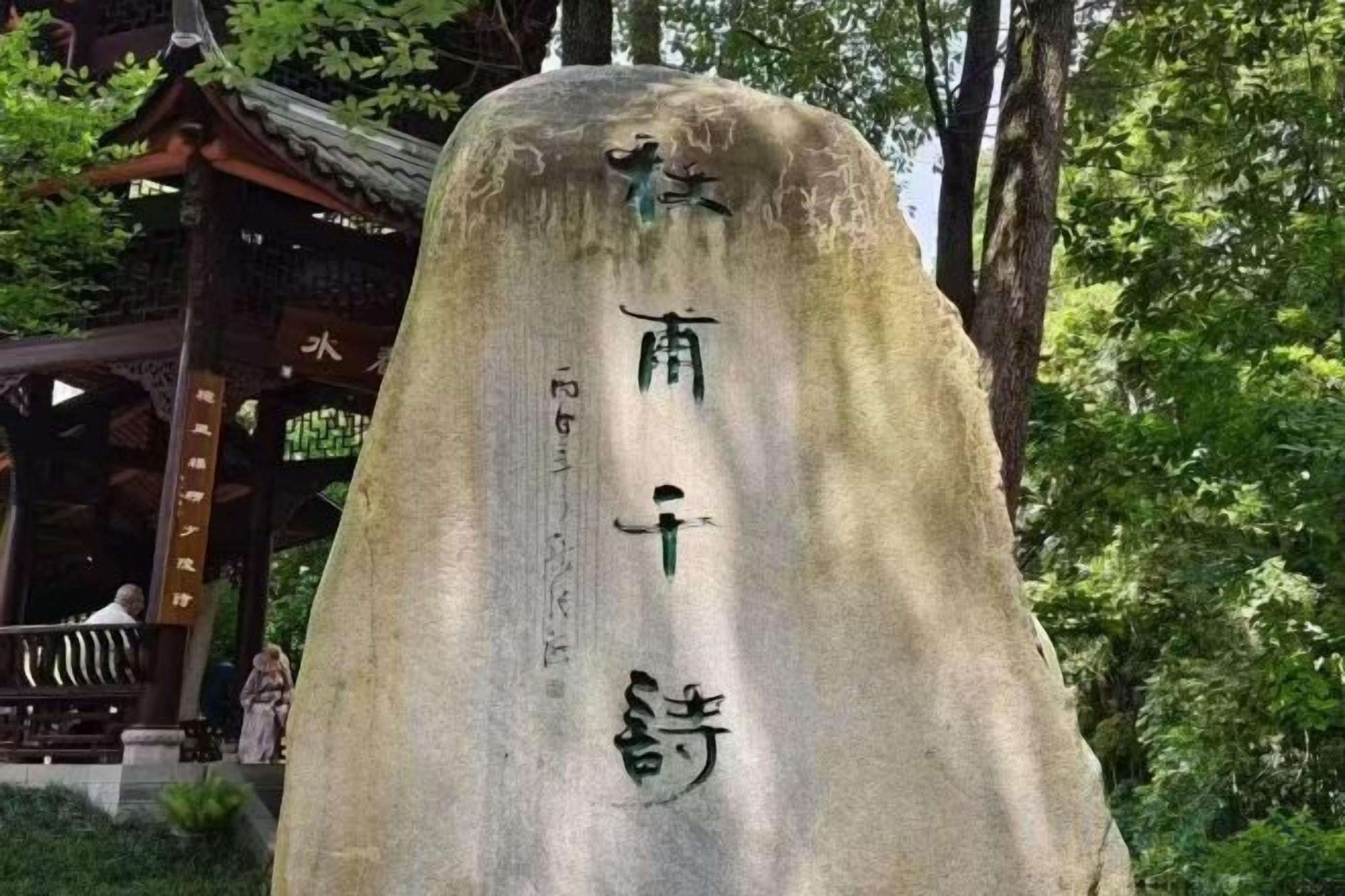 Un monumento in pietra con caratteri cinesi, circondato da alberi e un padiglione tradizionale.