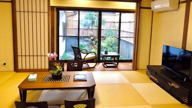 Een traditionele Japanse kamer met tatami vloerbedekking, een schuifdeur naar een tuin, een televisie en een klein tafeltje met een plant.