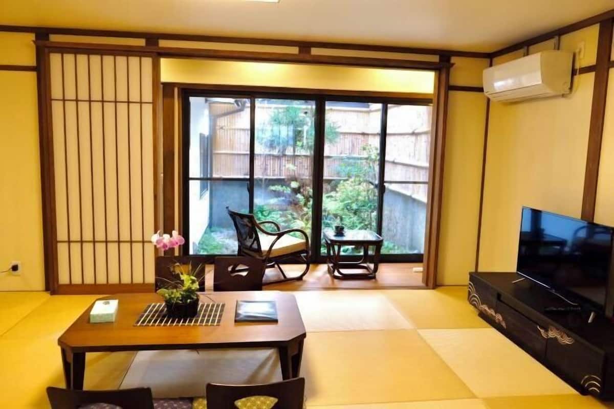 A traditional Japanese-style room with tatami flooring, a sliding door leading to a garden, a television, and a small table with a plant.