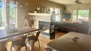 A modern kitchen with a stone fireplace, wooden dining table, and chairs. There is a balcony with a view of greenery. - Entire home in Palmer Lake, CO (Palmer Lake)