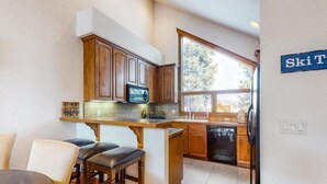 A modern kitchen with wooden cabinets, a built-in island with bar stools, and a large window offering a view of trees. - Ski In, Ski Out 3 Bedroom Townhome at Snowflake Lift - Private Hot Tub (Breckenridge)