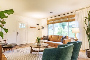 A living room with a sofa, two armchairs, a coffee table, and a lamp. There is a door with a coat rack, a large plant, and a window with curtains. - Pecan Belle Vintage Cottage | Fire Pit, Creek (Nashville)