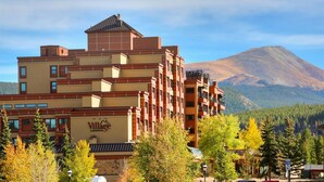 A multi-story hotel with a prominent Village sign, surrounded by trees and a mountain in the background. - Village at Breckenridge - 2 Bedroom Ski In/ski Out Condo (Breckenridge)