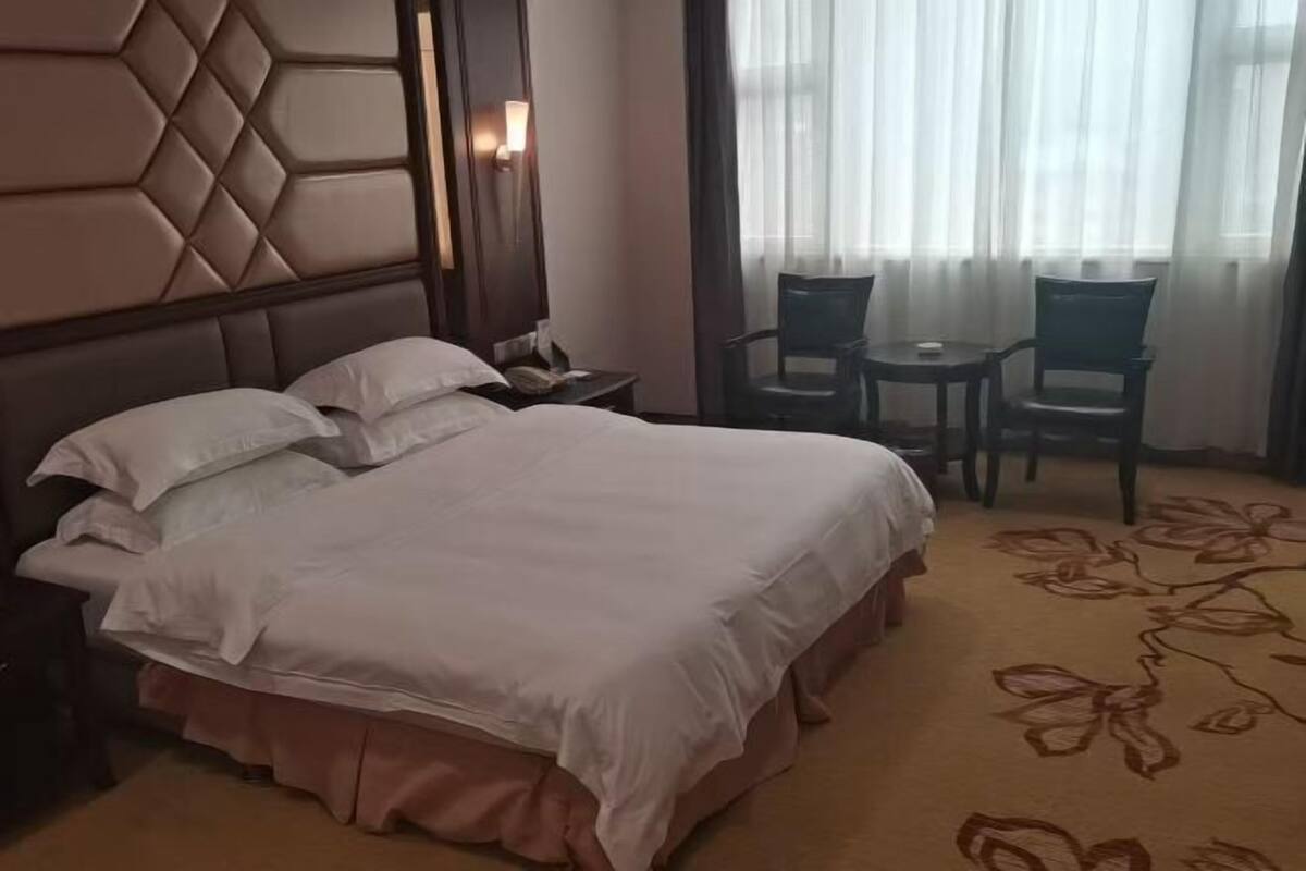 Een hotelkamer met een groot bed, een nachtkastje, twee stoelen en een raam met transparante gordijnen.